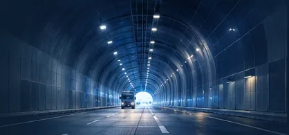 A lit road tunnel at night, seen along the carriageway, with a lorry in the distance and the arched lining lit by rows of lights.